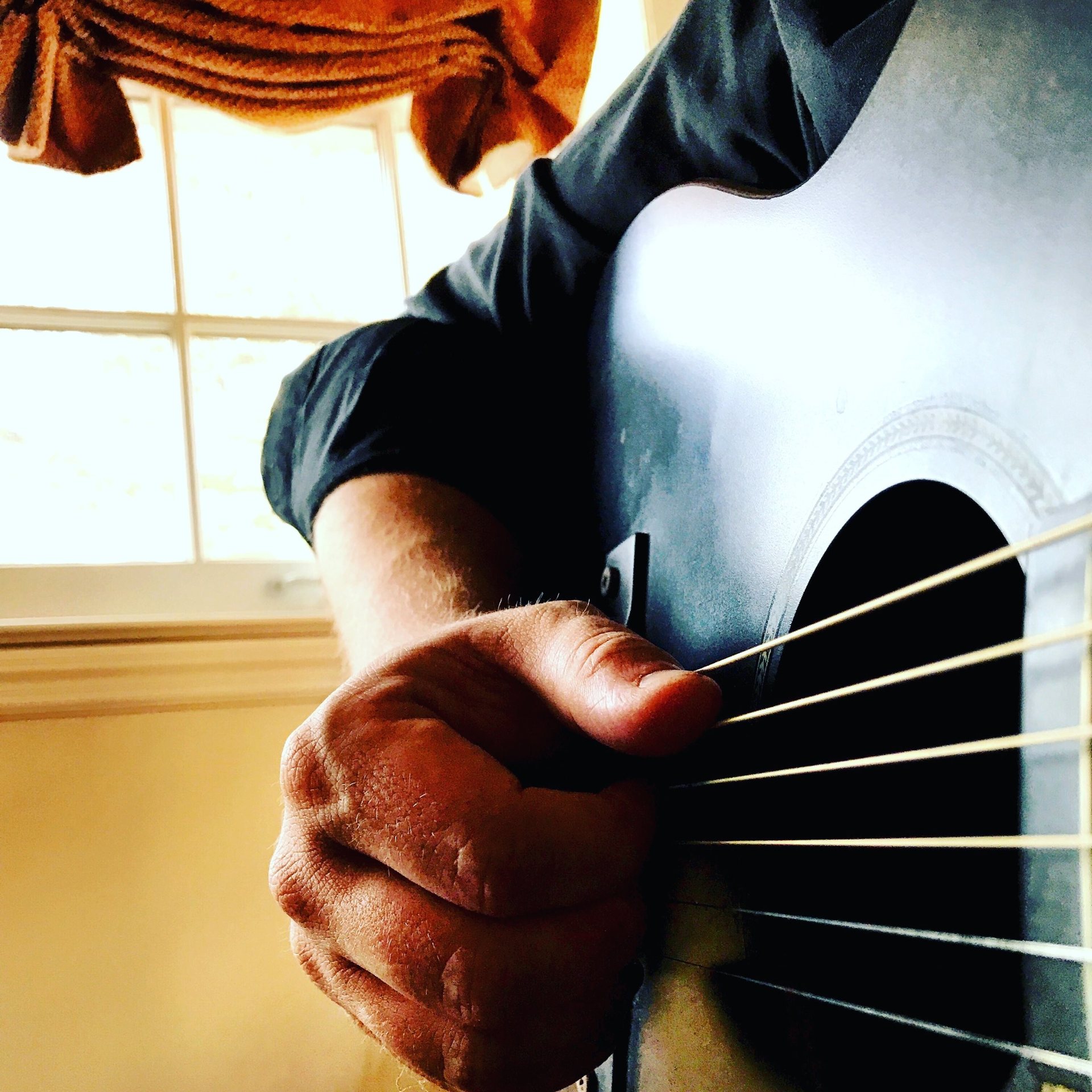 A hand picking the strings of a dark acoustic guitar beside a sunlit window
